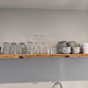 Op de afbeelding: Een houten plank toont diverse glazen, waaronder wijnglazen, bekers en koffiekopjes. Links staat een kleine potplant. De plank wordt ondersteund door zwarte metalen beugels en is bevestigd aan een lichtgrijze muur.