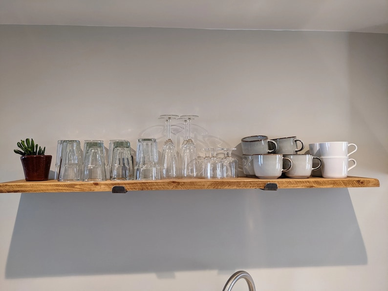 Op de afbeelding: Een houten plank toont diverse glazen, waaronder wijnglazen, bekers en koffiekopjes. Links staat een kleine potplant. De plank wordt ondersteund door zwarte metalen beugels en is bevestigd aan een lichtgrijze muur.