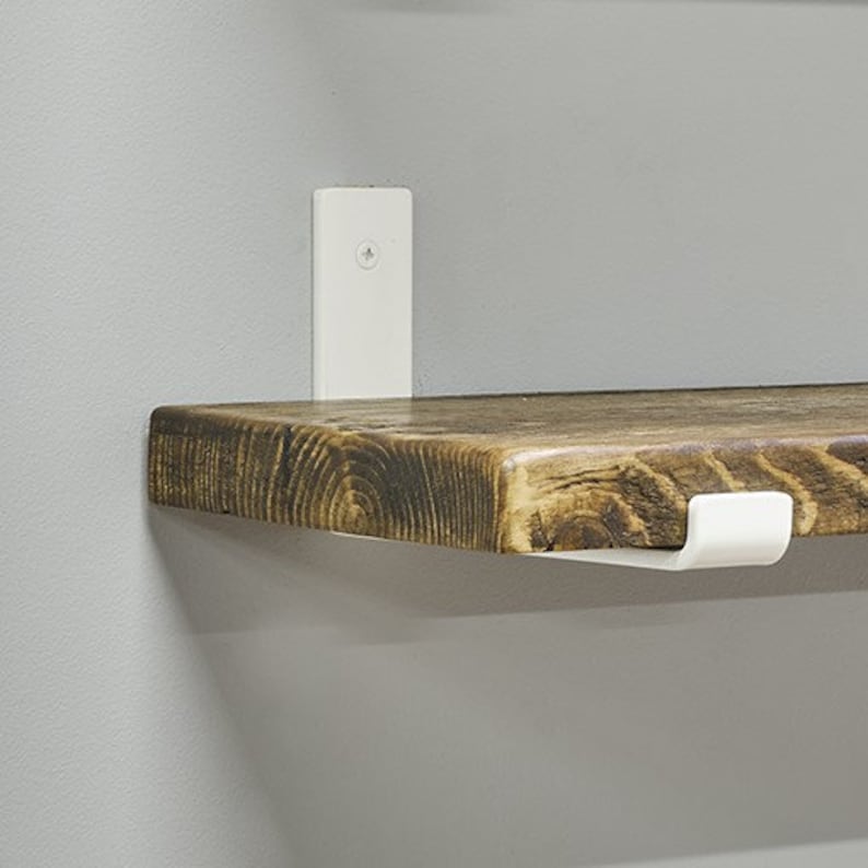 Op de afbeelding: Een rustiek houten plank met een donkere vlek, ondersteund door witte metalen beugels. De plank heeft zichtbare houtnerf en een natuurlijke rand, wat een warme uitstraling cre&euml;ert. De beugels zijn op een grijze muur gemonteerd.