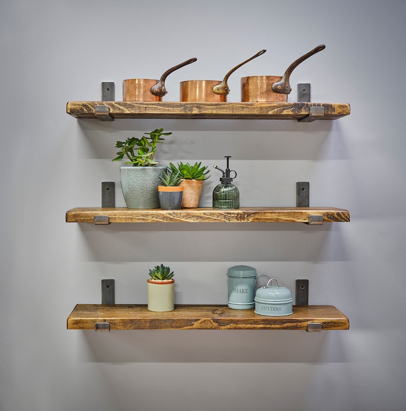 Op de afbeelding: Drie rustieke houten planken aan een grijze muur, elk ondersteund door donkere metalen beugels. De bovenste plank bevat koperen pannen, de middelste plank heeft potplanten en een groene glazen vernevelaar, en de onderste plank toont decoratieve blikken.