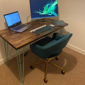 May include: A dark wood desk with teal hairpin legs and a teal velvet office chair with gold base and wheels. A laptop computer, a keyboard, and a book are on the desk. The book has the text "I survived another meeting without having to send an e-mail."