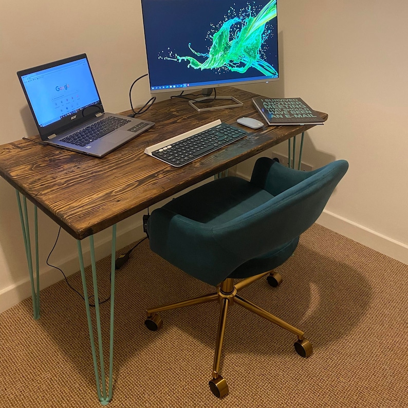May include: A dark wood desk with teal hairpin legs and a teal velvet office chair with gold base and wheels. A laptop computer, a keyboard, and a book are on the desk. The book has the text "I survived another meeting without having to send an e-mail."