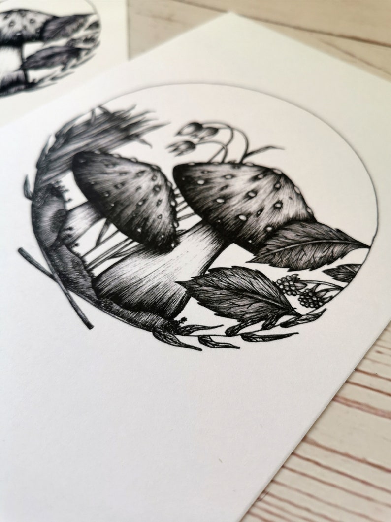 May include: Black and white ink drawing of two mushrooms with white stems and spotted caps surrounded by leaves and berries in a circle.