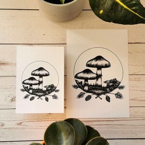 May include: Two black and white prints of mushrooms with pine cones and branches. The prints are on white card stock and are framed with a white border.