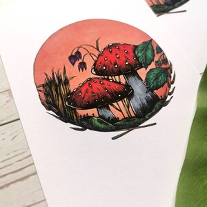 May include: A watercolor illustration of two red and white mushrooms with white spots in a forest setting. The mushrooms are surrounded by green leaves, tall grass, and a pink and orange sunset.