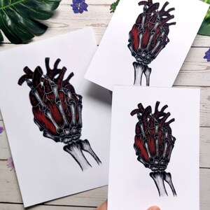 K&ouml;nnte beinhalten: Eine skelettartige Hand h&auml;lt ein rotes anatomisches Herz. Das Herz ist detailliert mit Venen und Arterien. Die Hand ist in einem realistischen Stil mit sichtbaren Knochen und Sehnen gezeichnet.