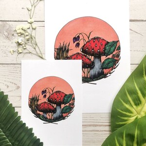 May include: Two red and white mushrooms with white spots, surrounded by green leaves and a pink background. The mushrooms are in a circle with a brown outline.