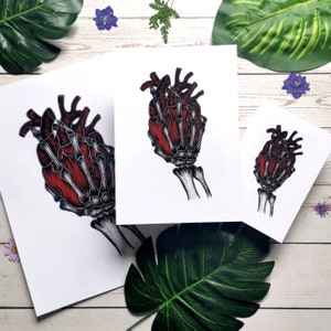 K&ouml;nnte beinhalten: Drei Drucke einer skelettartigen Hand, die ein rotes Herz h&auml;lt. Die Drucke sind auf wei&szlig;em Papier und in verschiedenen Gr&ouml;&szlig;en.