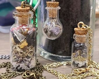 May include: Three glass vial necklaces with cork stoppers and metal chains. The vials contain various dried herbs, crystals, and other small items. The chains are in silver and gold tones, and the vials are displayed on a wooden surface.