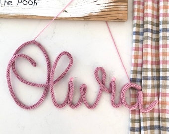 May include: Pink knitted yarn spelling the name "Olivia" hanging from a wooden sign with the quote "Forever, see." The Pooh written on it. The name is decorated with small pink beads.