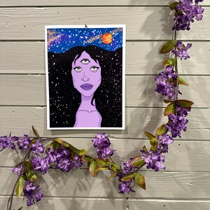 May include: A print of a woman with three eyes, purple skin, and long black hair. The background is a dark blue starry sky with a red planet and a ring.
