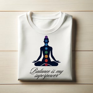 May include: White t-shirt with a black silhouette of a person in a yoga pose with seven colorful chakras. The text "Balance is my superpower" is written below the silhouette.
