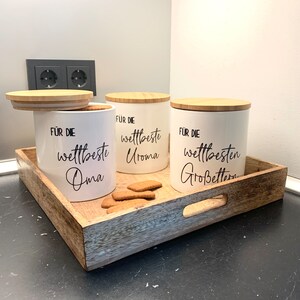 Könnte beinhalten: Drei weiße Keramikdosen mit Holzdeckeln. Jede Dose hat einen schwarzen Schriftzug: "Für die weltbeste Oma", "Für die weltbeste Uroma" und "Für die weltbesten Großeltern". Die Dosen stehen auf einem Holztablett mit einem Griff.