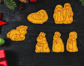 Puede incluir: Un conjunto de siete galletas de jengibre decoradas con una escena de la natividad. Las galletas son amarillas y presentan un bebé Jesús en un pesebre, tres reyes magos, una vaca, una oveja y María y José.