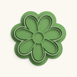 Pode incluir: Um cortador de biscoitos verde em forma de flor com um núcleo circular central e seis pétalas arredondadas. A ferramenta possui uma borda elevada para corte e uma base mais larga para manuseio, com um contorno simétrico e acabamento liso.