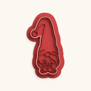 May include: A red plastic cookie cutter shaped like a gnome wearing a tall, pointed hat with a pom-pom at the tip. The gnome has a large nose, a long beard, and is holding a small, square gift box tied with a ribbon in its hands.