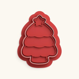 May include: A red plastic cookie cutter shaped like a stylized Christmas tree with three tiered branches and a five-pointed star at the top. The cutter features a thick outer frame and a thinner inner cutting edge for creating detailed shapes.