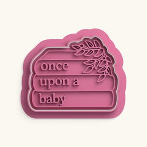 Puede incluir: Un cortador de galletas rosa con forma de pila de tres libros con el texto once upon a baby en relieve en la parte frontal. El libro superior presenta un diseño decorativo de rama con hojas en el lado derecho. Tiene un borde elevado.