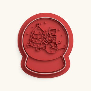 Puede incluir: Un cortador de galletas de plástico rojo con forma de bola de nieve. El diseño interior presenta un árbol de Navidad con una estrella en la punta y un muñeco de nieve con sombrero de copa y bufanda, grabado en el marco de plástico rojo.