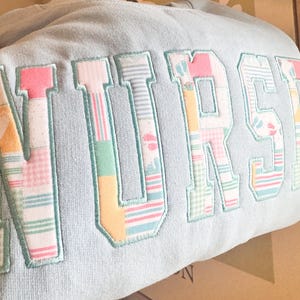 Puede incluir: Sudadera azul claro con la palabra "NURSE" bordada en letras grandes de patchwork colorido. Las letras presentan varios patrones pastel, incluyendo huellas de bebé, rayas y cuadros, con un contorno azul claro.