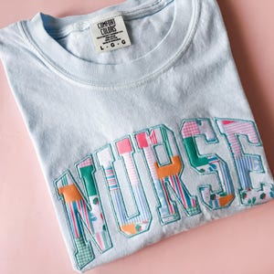 Puede incluir: Camiseta azul claro con la palabra "NURSE" bordada en tela de patchwork de colores.