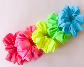 Könnte beinhalten: Ein Set aus vier Haargummis aus Stoff, die in einer diagonalen Linie angeordnet sind. Die Farben umfassen ein leuchtendes Pink, ein helles Neongelb, ein kräftiges Limettengrün und ein helles Himmelblau. Jeder Haargummi hat eine geraffte Textur.