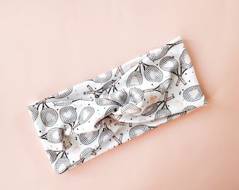 May include: White headband with a pattern of tennis rackets and balls. The headband is knotted on top.