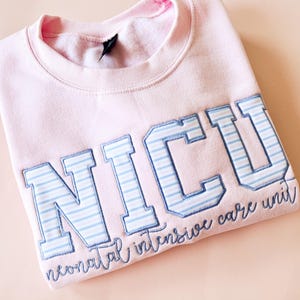 Puede incluir: Una sudadera rosa claro con las letras "NICU" en letras grandes a rayas azules y blancas. Las palabras "neonatal intensive care unit" están escritas en cursiva debajo.