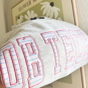Op de afbeelding: Een grijze sweatshirt met het woord "OBTECH" geborduurd in roze en lichtblauwe gestreepte letters. De sweatshirt is opgevouwen, met de tekst duidelijk zichtbaar. Een ingelijste print met de woorden "FLOWER MARKET" staat op de achtergrond.
