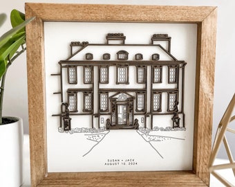 May include: A framed illustration of a house with a brown wooden frame. The illustration is in black and white and features a detailed drawing of a house with a front door and windows. The text "Susan + Jack August 10, 2024" is printed below the house.