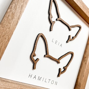 Puede incluir: Arte de pared enmarcado en madera con dos contornos de orejas de perro. Los nombres "LEIA" y "HAMILTON" est&aacute;n impresos debajo de las orejas. El marco es de madera marr&oacute;n claro y el fondo es blanco.