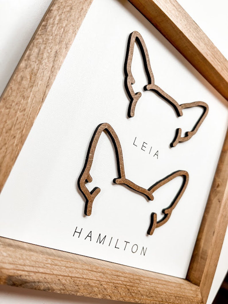 Puede incluir: Arte de pared enmarcado en madera con dos contornos de orejas de perro. Los nombres "LEIA" y "HAMILTON" est&aacute;n impresos debajo de las orejas. El marco es de madera marr&oacute;n claro y el fondo es blanco.