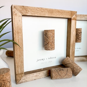 May include: A wooden frame with a white background and the text "jeremy + rebecca JULY 27, 2024". There are several wine corks attached to the frame.