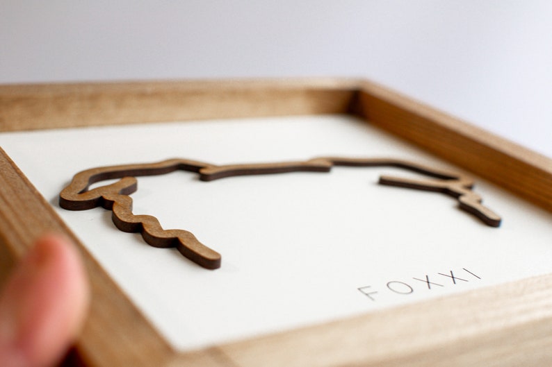 Puede incluir: Una obra de arte enmarcada en madera que presenta un contorno marr&oacute;n estilizado de una forma geogr&aacute;fica, posiblemente una costa o un r&iacute;o. La obra de arte est&aacute; sobre un fondo blanco, con la palabra "FOXXI" impresa en negro. El marco es de madera clara.