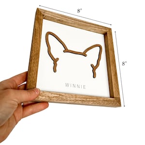Puede incluir: Letrero enmarcado de madera, 20x20 cm, con fondo blanco y contorno marr&oacute;n de orejas de perro. El nombre "Winnie" est&aacute; impreso en negro en la parte inferior. El marco es de madera marr&oacute;n claro.