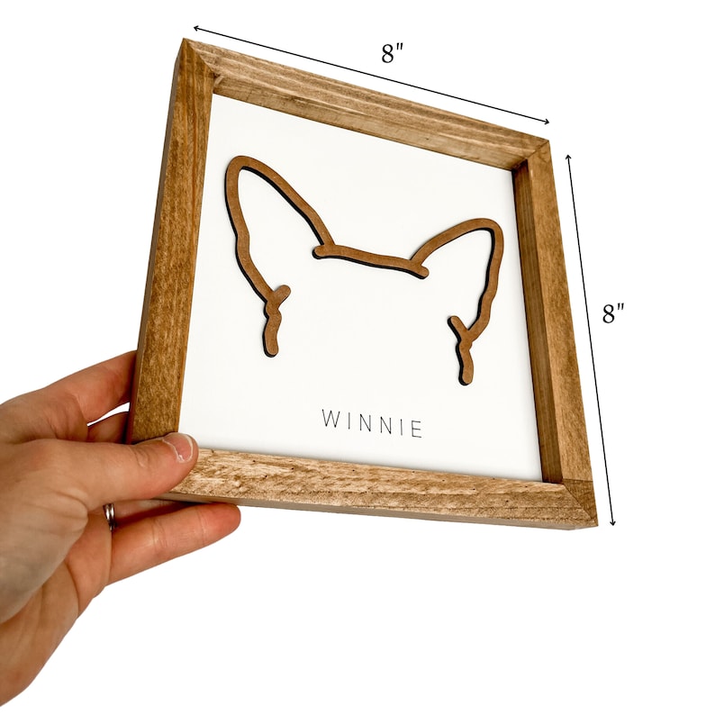 Puede incluir: Letrero enmarcado de madera, 20x20 cm, con fondo blanco y contorno marr&oacute;n de orejas de perro. El nombre "Winnie" est&aacute; impreso en negro en la parte inferior. El marco es de madera marr&oacute;n claro.