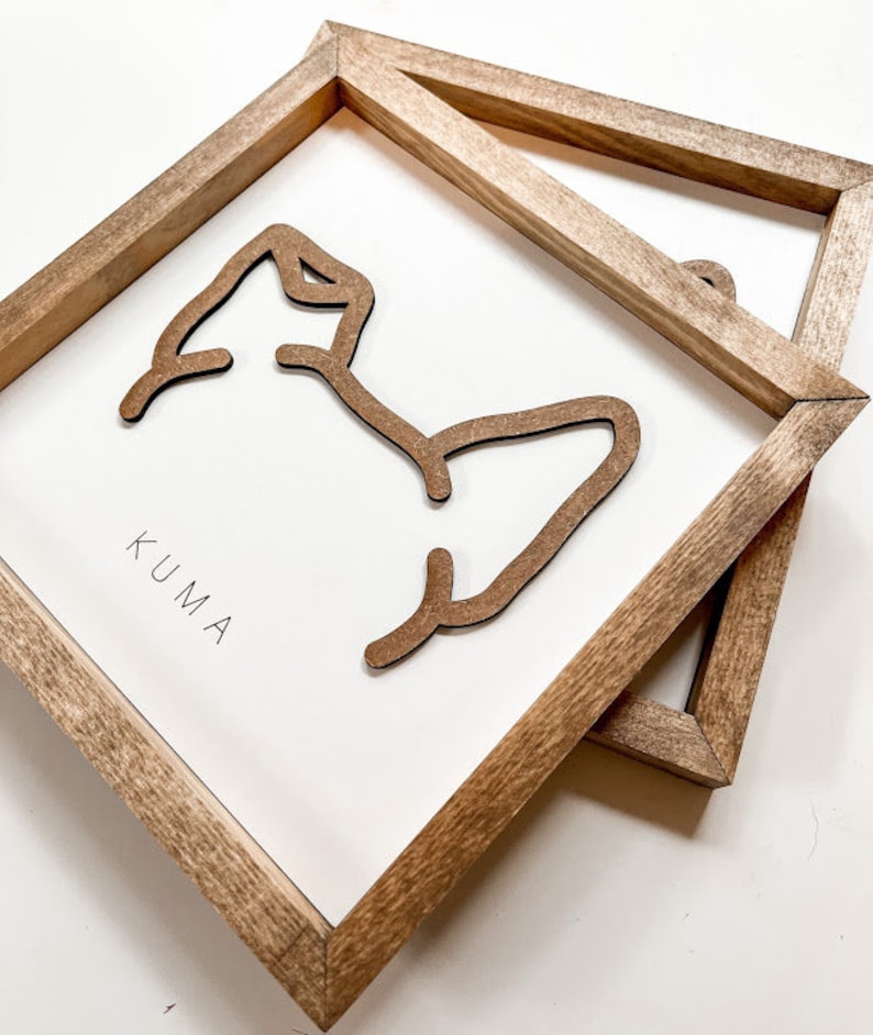 Puede incluir: Dos piezas de arte mural enmarcadas en madera. Cada pieza presenta un fondo blanco con un contorno marr&oacute;n de un oso y la palabra "KUMA" en negro. Los marcos son de madera marr&oacute;n claro.