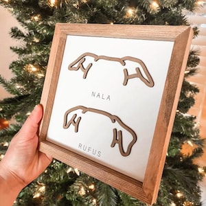 Puede incluir: Arte de pared enmarcado en madera con fondo blanco. El arte presenta dos contornos de orejas de perro marrones con los nombres "NALA" y "RUFUS" impresos debajo. El marco es de madera marr&oacute;n claro.