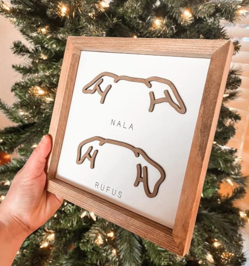 Puede incluir: Arte de pared enmarcado en madera con fondo blanco. El arte presenta dos contornos de orejas de perro marrones con los nombres "NALA" y "RUFUS" impresos debajo. El marco es de madera marr&oacute;n claro.