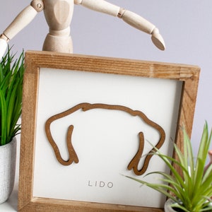 Puede incluir: Arte mural enmarcado en madera con una silueta de perro marr&oacute;n sobre un fondo blanco. La palabra "LIDO" est&aacute; impresa debajo. El marco es de madera marr&oacute;n claro. Plantas decorativas y un maniqu&iacute; de madera en el fondo.