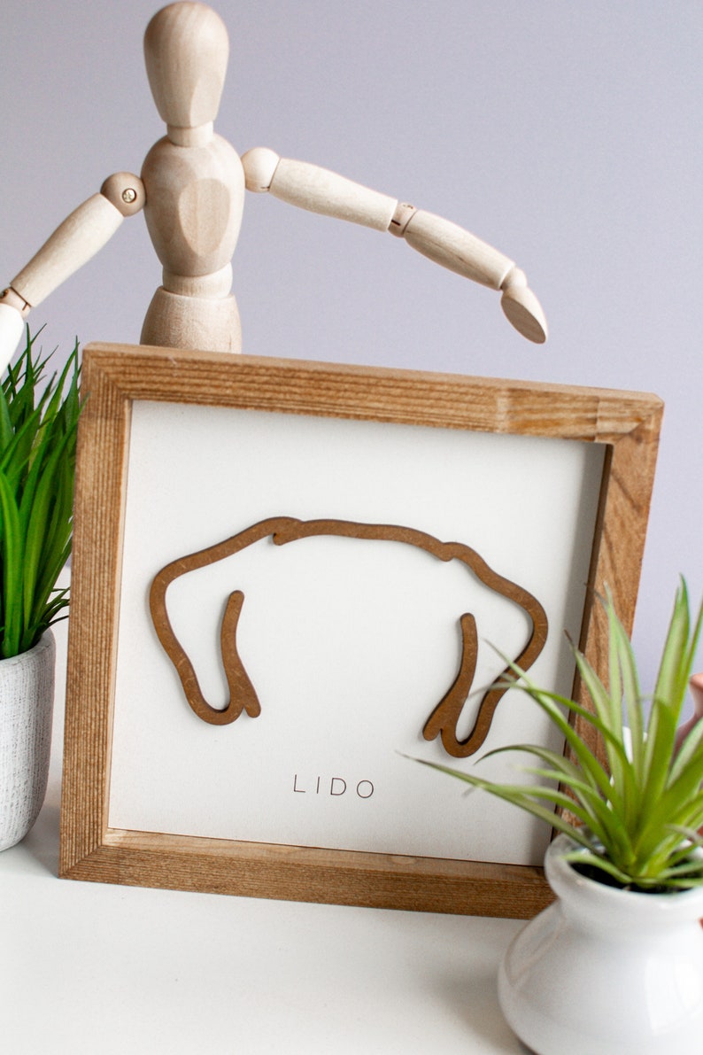 Puede incluir: Arte mural enmarcado en madera con una silueta de perro marr&oacute;n sobre un fondo blanco. La palabra "LIDO" est&aacute; impresa debajo. El marco es de madera marr&oacute;n claro. Plantas decorativas y un maniqu&iacute; de madera en el fondo.