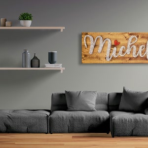 Può includere: Un cartello di legno con il nome "Michela" scritto con chiodi bianchi. Un piccolo cuore rosso è posizionato sopra la "i". Il cartello è appeso a una parete grigia sopra un divano grigio.