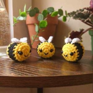 May include: Three crocheted bumblebees with black and yellow stripes, white wings, and black eyes. The bumblebees are sitting on a wooden table.