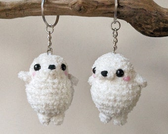 May include: Two white plush seal keychains with black eyes and pink cheeks. The keychains are hanging from silver chains.