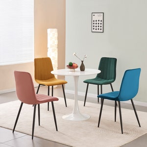 May include: Four velvet dining chairs in different colours: mustard yellow, teal green, dusty rose, and teal blue, surrounding a white round table with a pedestal base. The chairs have black metal legs.
