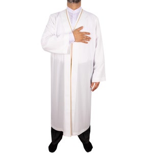 Puede incluir: Una t&uacute;nica blanca con ribete dorado, usada sobre una camisa blanca y pantalones negros. La t&uacute;nica tiene mangas largas y una parte delantera abotonada. La persona lleva una camisa blanca con cuello mandar&iacute;n. La persona est&aacute; de pie con la mano en el pecho.