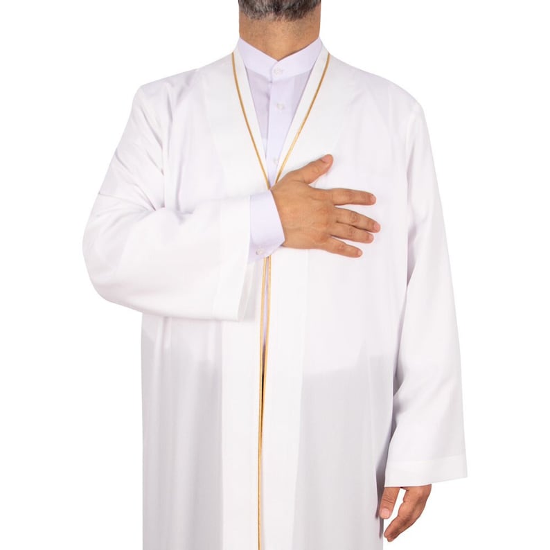 Puede incluir: Un thobe blanco, una prenda &aacute;rabe tradicional, con ribete dorado. El thobe se lleva sobre una camisa blanca con cuello. La persona se pone la mano en el pecho.