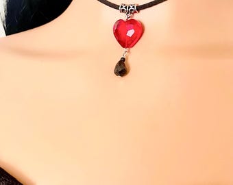 May include: A black choker necklace featuring a red heart-shaped pendant and a black teardrop-shaped charm. The heart pendant is faceted, catching the light. The necklace is worn around the neck.
