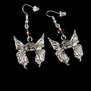May include: A pair of silver-toned butterfly earrings. Each earring features a detailed butterfly design with intricate wing patterns. A small purple bead is suspended between the butterfly and the ear hook. The earrings are displayed against a black background.