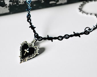 Könnte beinhalten: Eine Halskette in Schwarz und Silber mit einem herzförmigen Anhänger. Der Anhänger hat ein Kreuz und einen dekorativen Rand. Die Halskette hat ein Stacheldraht-Design mit blauen Akzenten in der Nähe des Verschlusses.